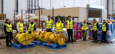 Nine people in yellow hi-vis jackets stand in a loose line, looking at the camera, surrounded by pallets of equipment
