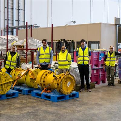 Nine people in yellow hi-vis jackets stand in a loose line, looking at the camera, surrounded by pallets of equipment