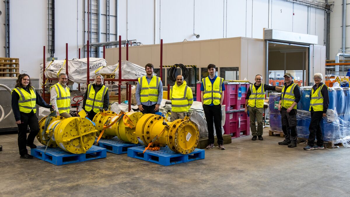 Nine people in yellow hi-vis jackets stand in a loose line, looking at the camera, surrounded by pallets of equipment