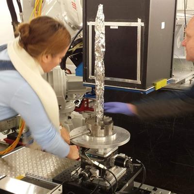 Two researchers positioning a long, thin sample wrapped in foil in a steel clamp