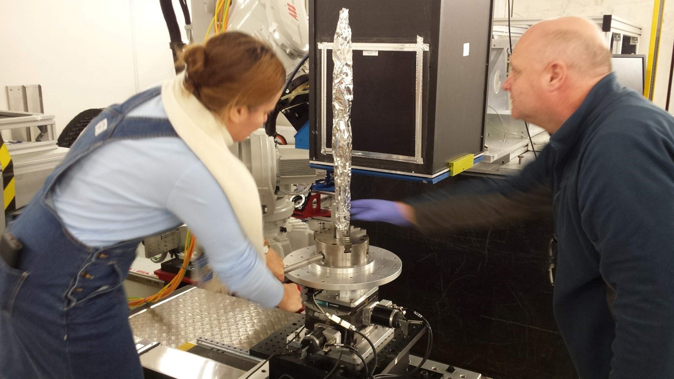 Two researchers positioning a long, thin sample wrapped in foil in a steel clamp