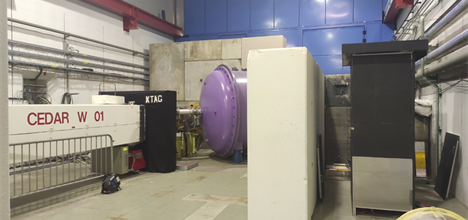 A large concrete room containing the NA62 experiment in white, black and purple cases and, to the right, a tall black cupboard