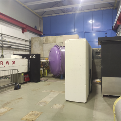 A large concrete room containing the NA62 experiment in white, black and purple cases and, to the right, a tall black cupboard