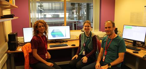 Three people, sat in an office around a computer. The computer screen displays a large colourful graph