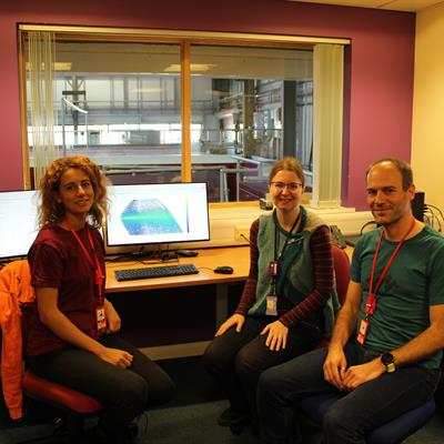 Three people, sat in an office around a computer. The computer screen displays a large colourful graph