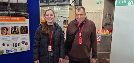 Two people stand smiling at the camera, inside ISIS TS1. To one side are posters describing ISIS research