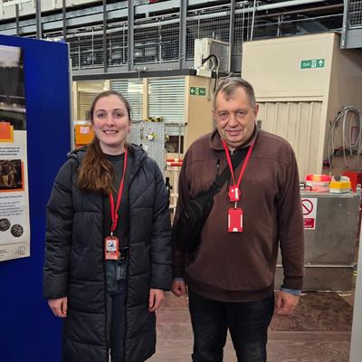 Two people stand smiling at the camera, inside ISIS TS1. To one side are posters describing ISIS research