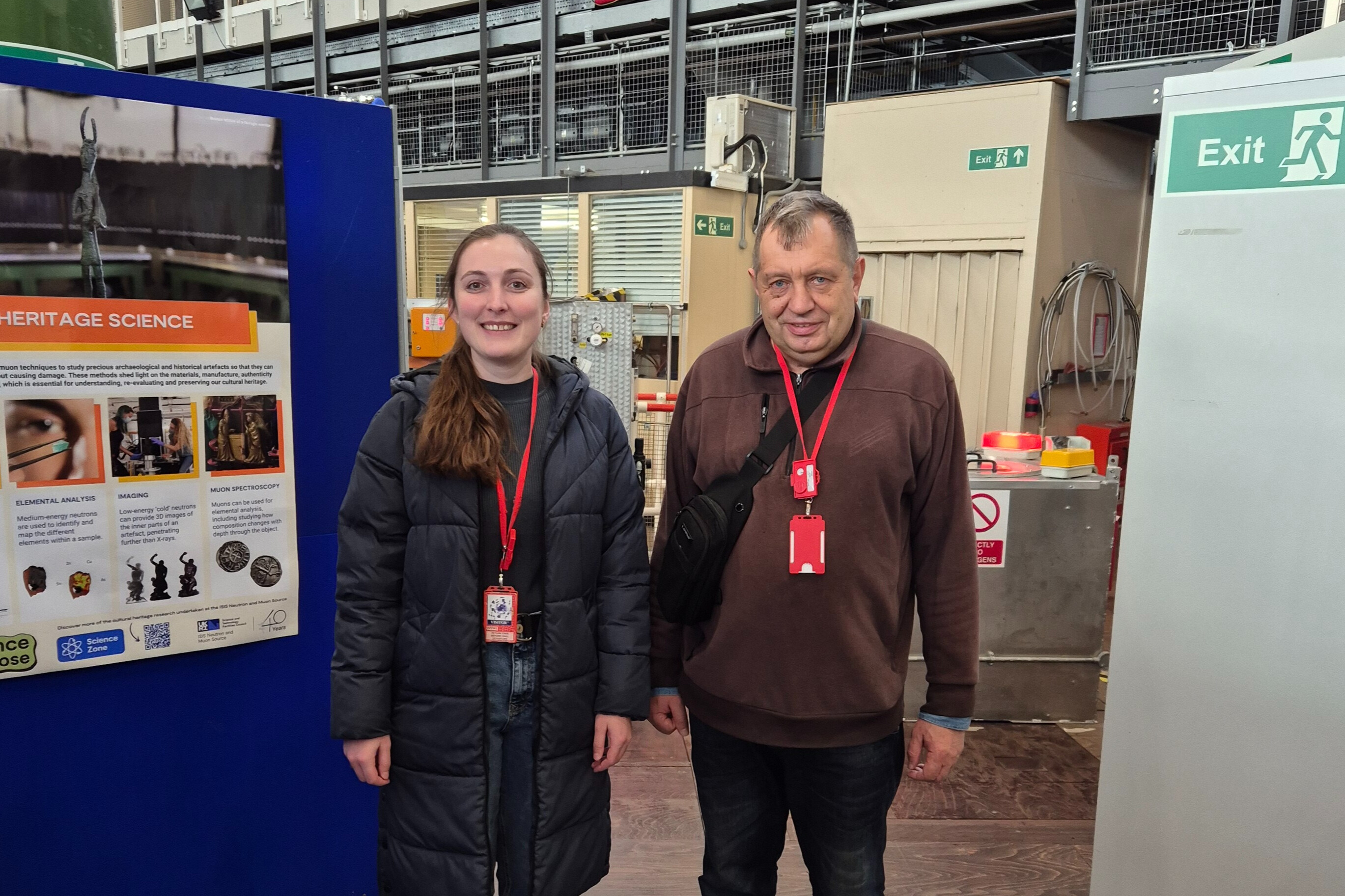 Two people stand smiling at the camera, inside ISIS TS1. To one side are posters describing ISIS research