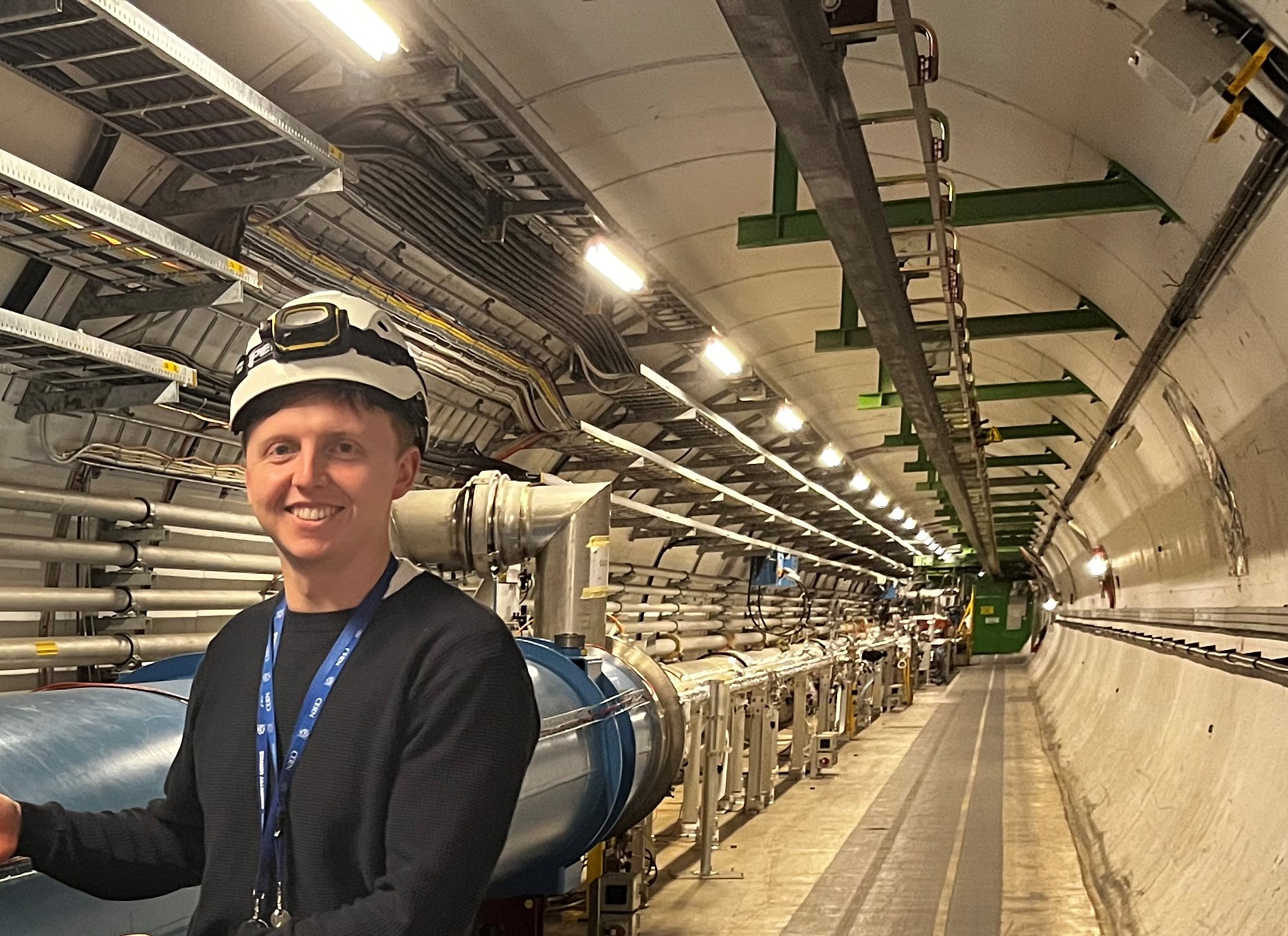 a man stood in front of a large underground apparatus, wearing a hard hat and smiling at the camera