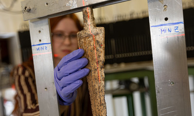 A close up shot of an old sword with a gloved hand touching it. A woman can be seen in the background, slightly blurred.